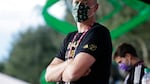 Portland Timbers team owner Merritt Paulson stands on the sidelines of a soccer pitch, wearing a mask with small Timbers logos.