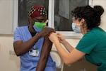 A woman in blue gown wearing mask receives vaccination from another woman.