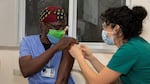 A woman in blue gown wearing mask receives vaccination from another woman.