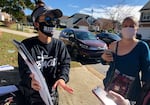 Two canvassers stand on a sidewalk talking with a person off camera who is holding a cellphone. One canvasser is wearing a Biden face mask, a visor and sunglasses while gesturing to informational placards she's holding. The other holds her phone in her right hand as she listens.