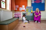 A 4-year-old girl kneels on the floor in her kindergarten classroom as her teacher sits in a small wooden chair beside her. Toy cars are scattered on the tile floor.