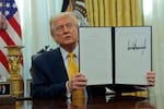 In this photo, President Trump holds up an executive action he signed. He is seated at a desk in the Oval Office and is wearing a dark blue suit and golden-colored tie.