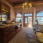 Pictured is the Star Saloon, a western-style bar with an ornate wood work. A Chandelier hangs above and four people are shown aiding in restoration efforts. Two people in blue shorts are painting a back wall with a French-style door entrance. Two others, one in white and one in gray, are working at the bar area.