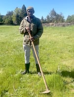 An African man wearing tall boots, grey pants, and a green jacket and beanie, stands in a field smiling, holding a garden hoe.