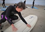 A woman on the beach crouches over a surfboard and applies wax to its topside.