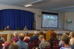 A roomful of people sit in chairs facing a panel of state officials and a video screen displaying a presentation.