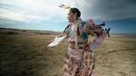 A member of the Umatilla Tribe performs a powwow dance in jingle regalia outside in a field.