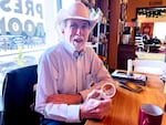 Jeff Poppe looks at the camera and holds a magnifying glass. He's wearing a striped shirt and cowboy hat.  He's pictured in a coffee shop.