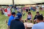 A group of boys and men bang a drum in a circle as dancers go by in the background. Among the dancers is a woman in bright red regalia in mid-spin.