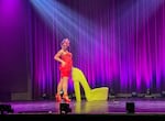 A woman poses on stage in front of purple  and blue curtains. She's wearing a red dress, hat, and heels with a bow, posing beside a big high-heel-shaped chair.