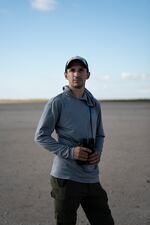 Justin LeClaire, a conservation biologist, holds a pair of binoculars while standing on the sand flats. He's wearing a baseball cap and a hoodie.