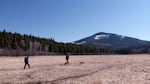 Two people carrying backpacks walk across a meadow with a dog. Forest and a mountain are in the background.