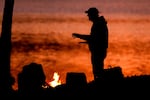 A person stands by a campfire with a lake or river in the background.