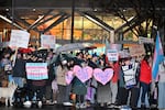 A large gathering of people are shown, most carrying homemade signs, in front of a hospital building. It is raining and people have umbrellas open. 