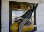 A person wearing a black beanie, black sweatshirt and gray pants rides a mechanical corn dog.