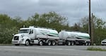 A white fuel truck marked "Christensen" pulls a trailer with a second fuel tank on a paved road lined with leafy green trees.