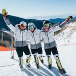 Three women stand on a snowy mountain on their skis, close to each other arm-in-arm, smiling and waving at the camera. They all have matching outfits, including matching high-tech shorts.