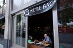 Two people sitting next to a large open window at a restaurant are photographed from outside. Above them, a banner reads "Q we're back."