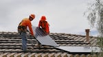 In this July 28, 2015, photo, electricians Adam Hall, right, and Steven Gabert, install solar panels on a roof for Arizona Public Service company in Goodyear, Ariz. Traditional power companies are getting into small-scale solar energy and competing for space. The emerging competition comes as utilities and smaller solar installers fight over the future of the U.S. energy system.  