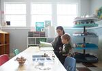 Sitting at a table in a preschool classroom, a mother reads to her toddler son.