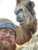 A bearded white man wearing glasses and a gray beanie poses for a selfie with his pet brown camel.