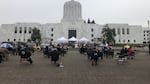 People sit outside in chairs on a gray day facing the Oregon State Capitol building for an event.