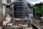 A man wearing rubber boots and holding a shovel stands in front of a water tank with a hose on it.