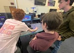 Three boys are gathered around a computer screen: on the left, one in a pink shirt is directly working on the computer, in the middle, another in a burgundy hoodie, and on the right, another stands in an army green hoodie.