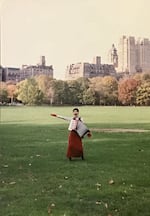 A person stands in a grassy field wearing an accordion across her chest, with tall urban buildings in the background.