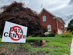 This photo shows a white yard sign that says in all caps, "NO DATA CENTER!" A red ring with a slash through it appears on top of the text. Stretching behind the sign is a grassy green yard, and in the background stands a brick house.