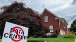 This photo shows a white yard sign that says in all caps, "NO DATA CENTER!" A red ring with a slash through it appears on top of the text. Stretching behind the sign is a grassy green yard, and in the background stands a brick house.