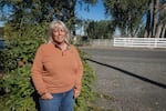 A blonde woman in an orange fleece pullover and blue jeans in front of a some plants and a rural road.