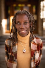 A woman wears a yellow turtle neck beneath a plaid jacket, with a pendant necklace around her neck. She smiles slightly as she looks at the camera.