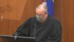 A judge, wearing a face mask, glasses and in his judicial robes sits on the bench talking into a small microphone as he addresses the court. A reflection of some equipment is visible in the plexiglass divider in front of him.