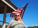 A woman in a sleeveless top stands in front of an American flag that hangs against a bright blue sky.