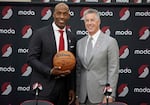 Two individuals stand side by side with one holding a basketball in front of a black background with the Portland Trail Blazers pinwheel logo and the word "moda" emblazoned all over it.