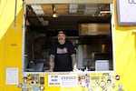 The chef and owner of a yellow food cart stands inside, looking to camera.