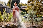 Brenda Jacobs does garden maintenance at Daybreak Cohousing in Portland. She is a woman with short, red hair wearing overalls. She's watering plants with a hose.