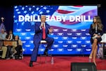 In this photo, Donald Trump sits on a chair on a stage while holding a microphone. Behind him is a screen that says "Drill baby drill" in large letters.