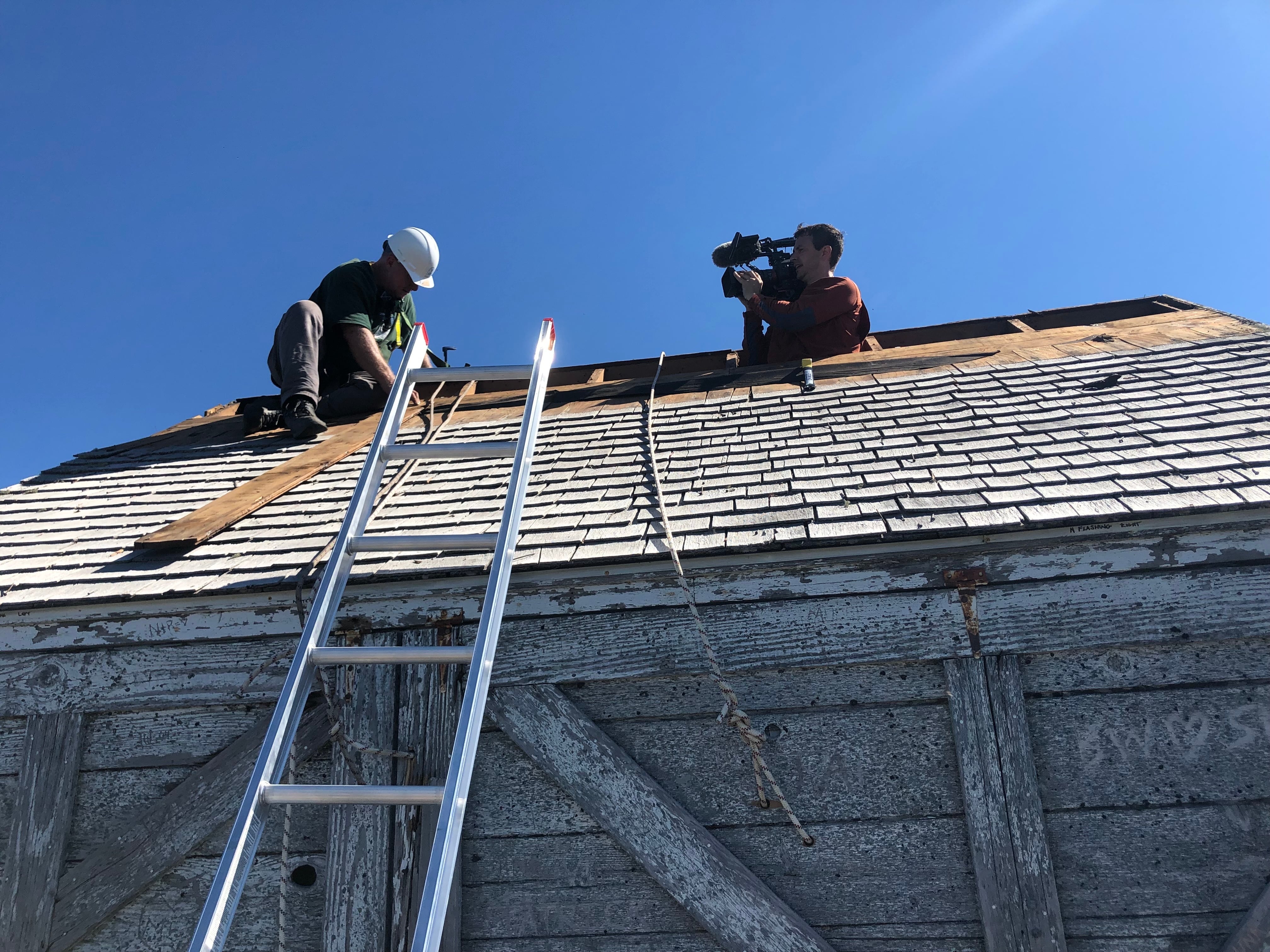 Don Allen disassembles the roof of the historic High Rock lookout in 2021, carefully removing and saving each cedar shingle to be put back after the restoration. OPB videographer Brandon Swanson films Allen for