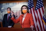 A masked Nancy Pelosi addresses the media from a lectern in the Capitol as a masked Blumenthal looks on from over her right shoulder. American flags hang in the background.