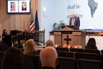 Images of Saylor Graham, 7, and Becky Drury are displayed on the screen as Pastor Richard Peil speaks during a service at Victory Baptist Church, Oct. 18, 2026, in Bend, Ore., to honor the victims of the recent shooting at Silver Rail Elementary School.