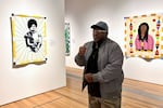 Artist Gary Tyler stands before his quilt, "Defiant, 1976," (2023) created in 2023. Tyler says the gesture he iis making with his hands was intended to inspire hope and resistance for the people who were protesting the circumstances of his trial.
