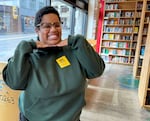 Katherine is a Black woman with short hair and glasses, and she's posing with both hands under her chin and smiling. The window out to the street and shelves of books are in the background.