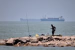A man stands beside a fishing pole along the shore as cargo ships and commercial vessels are seen in the Strait of Hormuz off Bandar Abbas, Iran, Wednesday, June 17, 2026.