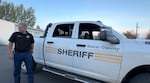 A sheriff stands in front of a patrol truck with a holding cell in the back.