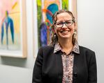 A smiling woman in blue glasses, a patterned shirt and black blazer. A row of paintings hangs on the wall in the background.