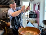A white man wearing an Oregon State Beavers baseball cap, a light blue short-sleeved collared shirt and a black apron  attach a whisk of clear corn syrup to a hook above a cooper kettle in a candy shop kitchen.