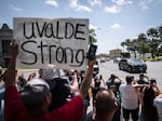 People stand together on the side of a road watching a motorcade drives by. One person holds a sign that reads "Uvalde strong"
