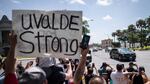 People stand together on the side of a road watching a motorcade drives by. One person holds a sign that reads "Uvalde strong"